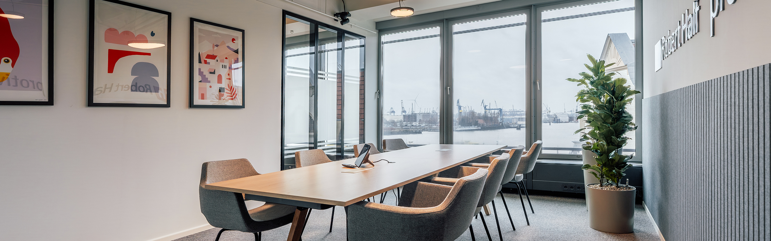 Modern meeting room with a large wooden table and upholstered chairs, floor-to-ceiling windows overlooking the Port of Hamburg, glass walls, wall graphics and a plant as a green accent.