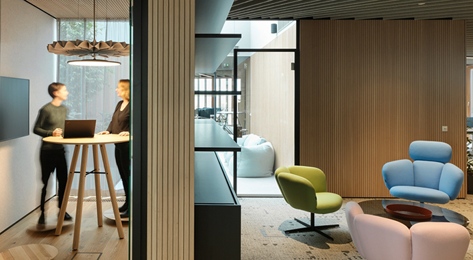 Moderner Bürobereich mit Besprechungsraum und Loungebereich, in dem zwei Personen an einem Stehtisch arbeiten und farbige Sitzmöbel flexible Nutzung ermöglichen.