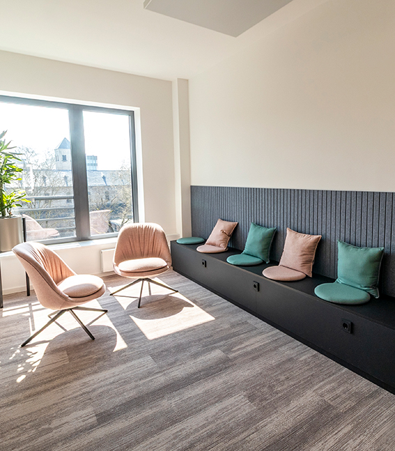 Quiet office retreat area with a cushioned bench, armchairs and pillows in front of a large window for focused work or relaxation.