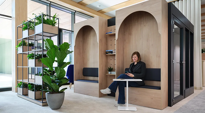 Modern office space with wooden alcoves and built-in benches; a woman sits at a small table looking at her smartphone; plant shelf next to her.