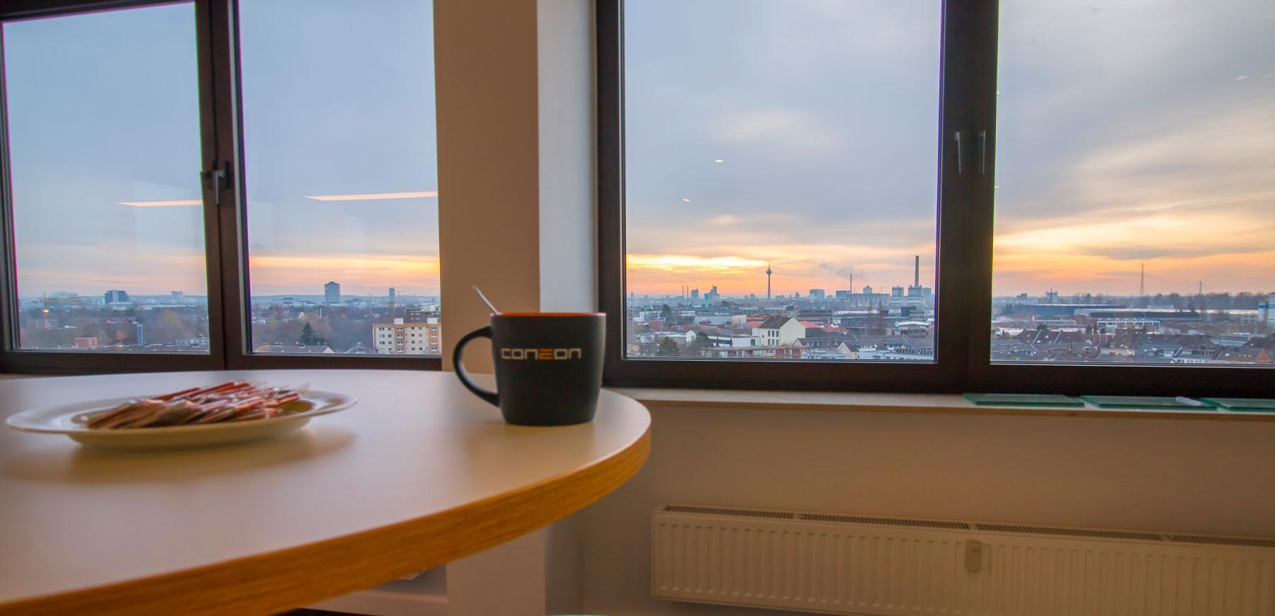 Eine Coneon-Tasse und ein weißer Teller auf einem Holztisch mit Fenstern und Blick auf die Stadt im Hintergrund.