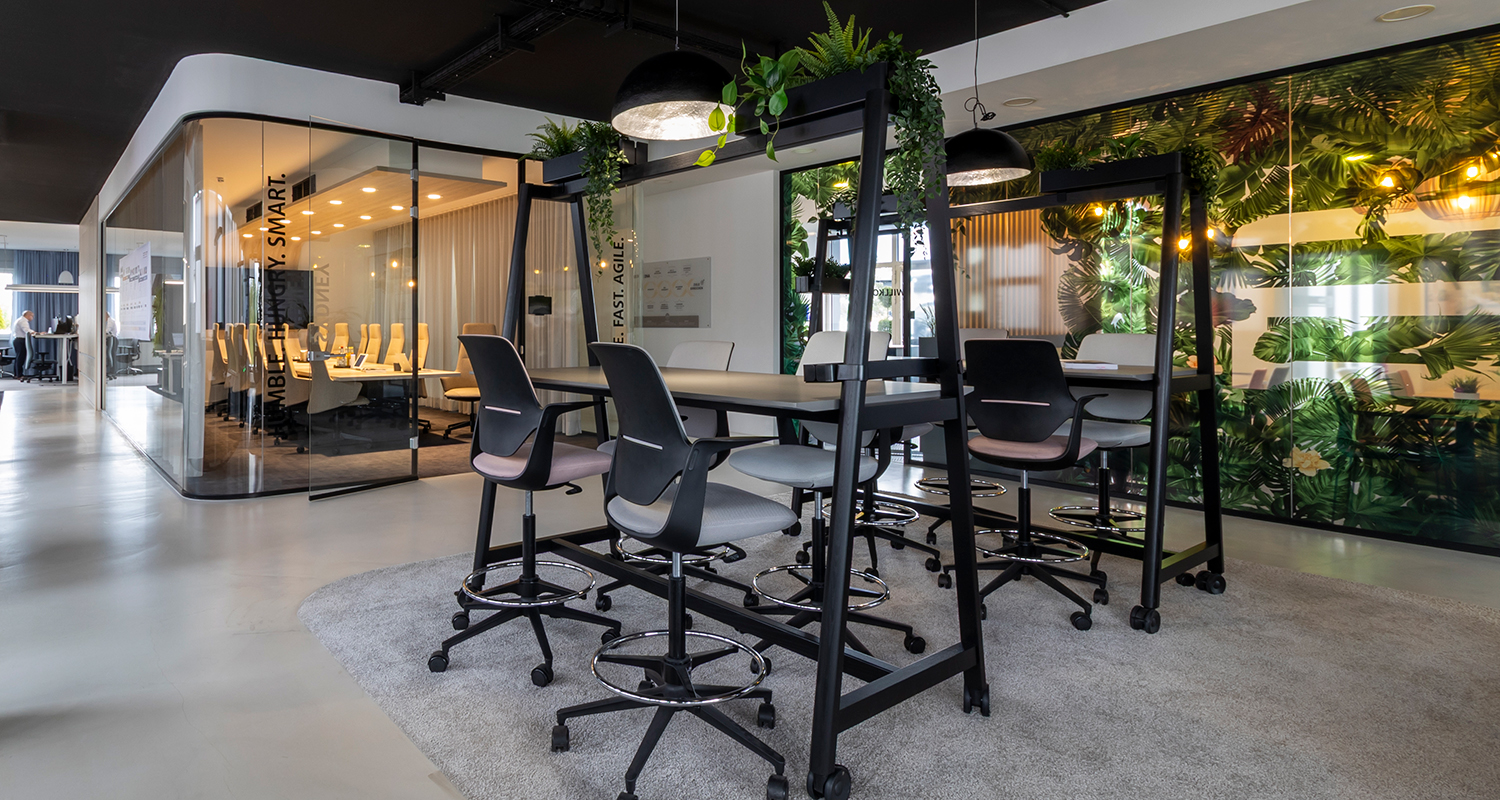 Open-plan office area with standing table and high swivel chairs, black ceiling and pendant lights; in the background, a glass-walled meeting room and a wall with a tropical plant motif.